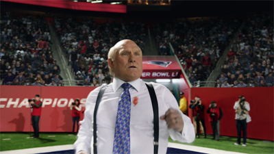 A bald man in a white dress shirt and blue patterned tie stands in a crowded sports stadium.