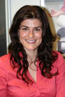 A smiling woman with long, wavy dark hair wearing a bright pink button-down shirt.