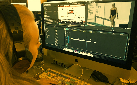 A person with headphones looks at a computer monitor displaying video editing software and footage.