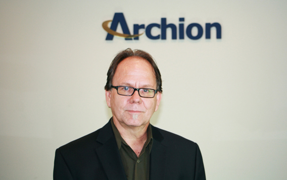 A man with glasses and a dark blazer stands before a white wall with the blue Archion logo.