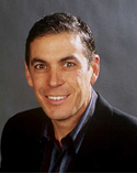 A smiling man with short dark hair wearing a dark blazer and a collared shirt.