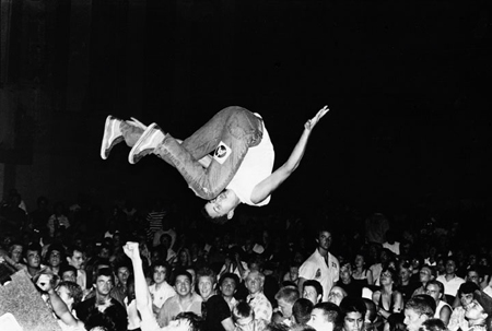 A person in a white shirt and jeans performs a backflip mid-air above a large crowd at night.
