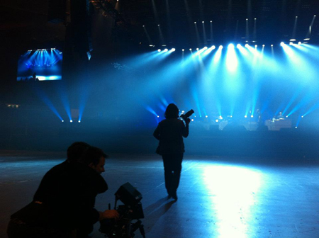A silhouette of a camera operator filming a brightly lit concert stage with blue spotlights.