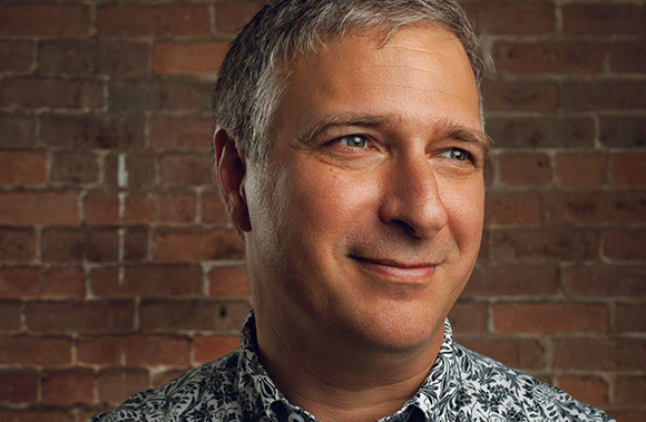 A man with graying hair and a patterned shirt smiles while looking off-camera against a brick wall.