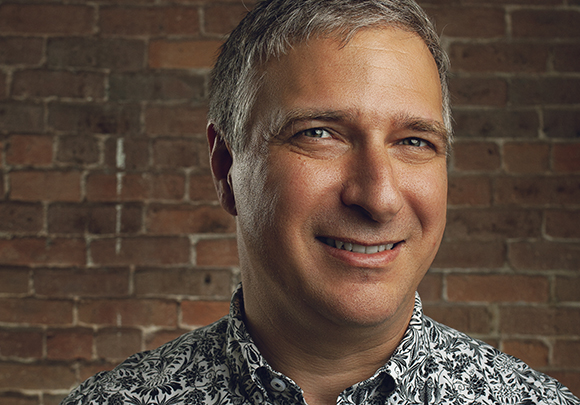 A middle-aged man with short graying hair smiling against a brown brick wall.
