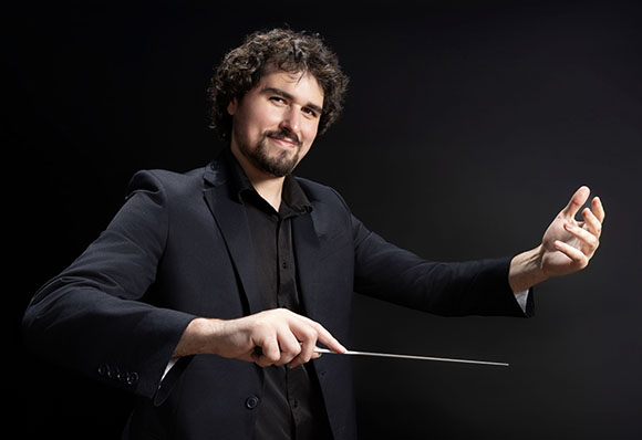 A man with curly hair and a beard wearing a black suit, holding a conductor's baton against a dark background.