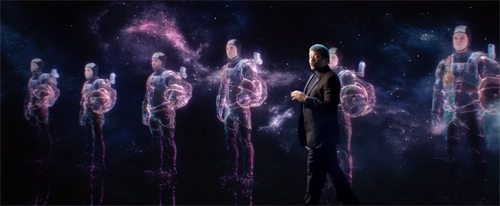 A man in a dark suit walks across a stage in front of multiple glowing, holographic figures in space suits.
