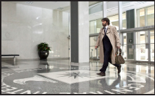 A man in a tan trench coat and suit walks through a bright, modern lobby with marble floors.