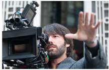 A man with a beard looks through the viewfinder of a large professional film camera while holding up a hand.