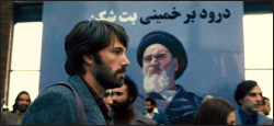 A bearded man in profile stands before a large blue poster featuring a portrait of Ayatollah Khomeini.