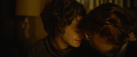Two women with wavy hair leaning their foreheads together in warm, dim light.