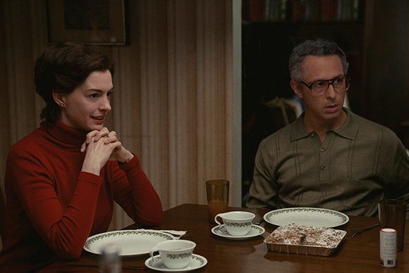 A woman in a red turtleneck looks toward a man in a green polo shirt at a dining table.