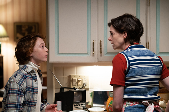 A person in a plaid shirt looks at another person in a striped vest in a kitchen setting.