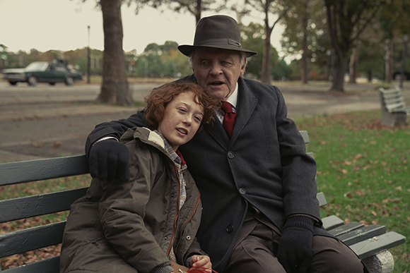 An older man in a hat and coat sits on a park bench with his arm around a young girl with red hair.