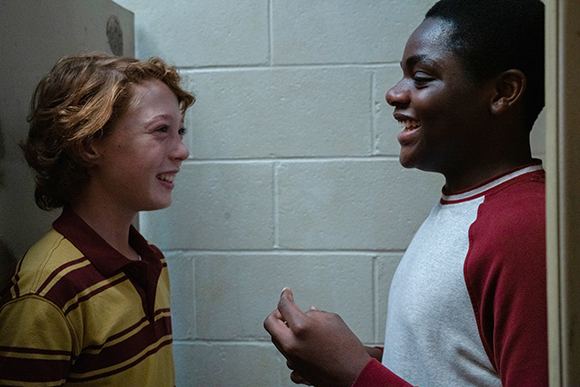 A young boy with curly red hair and a boy with dark skin smile at each other in a hallway.