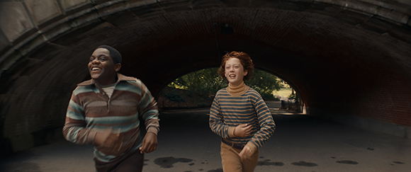 A young Black boy and a young white girl with red hair run joyfully through a dark brick archway.