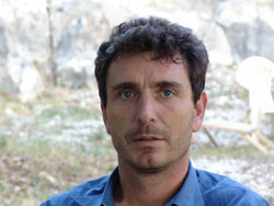A man with dark, wavy hair and blue eyes wearing a blue collared shirt outdoors.
