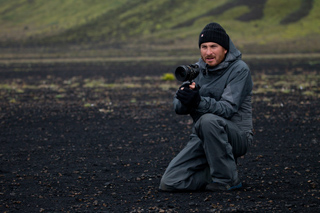 A man in a grey jacket and black beanie kneels on dark volcanic soil while holding a professional camera.