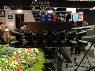 A camera rig with multiple lenses is mounted on a tripod, with a Hitachi display in the background.