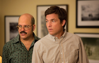 A man with glasses and a mustache stands next to a man in a checkered shirt, both looking surprised.
