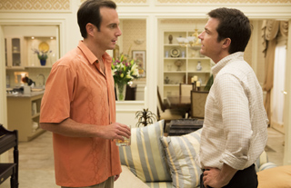Two men in button-down shirts stand facing each other in a brightly lit, upscale living room.