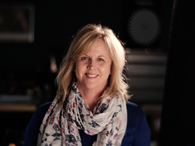 A smiling blonde woman wearing a blue shirt and a white floral patterned scarf.