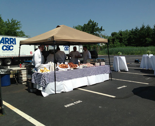 Caterers preparing food at a long table under a tan canopy in a parking lot.