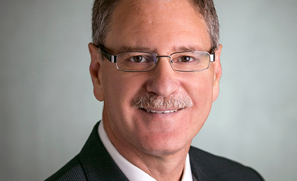A smiling middle-aged man with short brown hair, glasses, and a gray mustache wearing a dark suit.