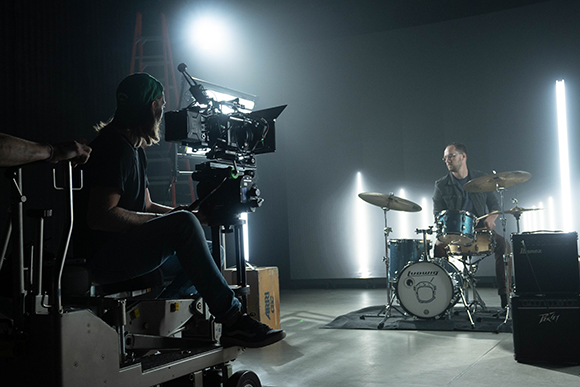 A camera operator on a rig films a man playing a drum kit in a dark studio with bright spotlights.