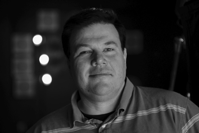 Black and white close-up portrait of a man smiling slightly against a dark, blurred background.