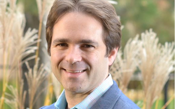 A man with brown hair smiling at the camera, wearing a light blue patterned shirt and blue blazer.
