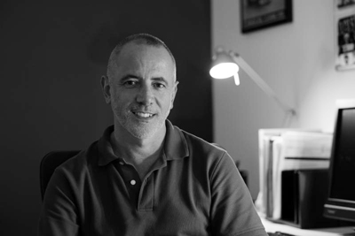 A man in a polo shirt smiles at the camera in a dimly lit office setting.