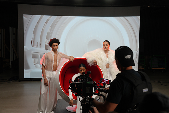 Models in white clothing pose around a large red ring in a studio with a digital backdrop and a camera operator.