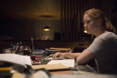 A woman with reddish-blonde hair sits at a cluttered desk in a dark room, looking intently forward.