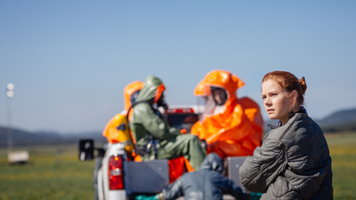 A young woman with red hair looks toward people in orange and green hazmat suits in a field.
