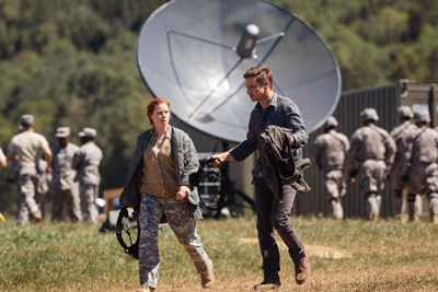A woman in camouflage and a man in a dark shirt walk through a field near a large satellite dish.
