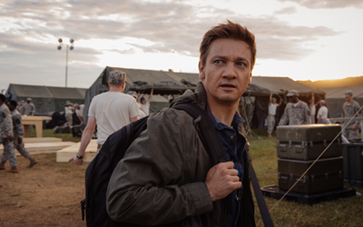A man wearing a dark jacket and backpack looks back toward a military camp with tents and personnel.