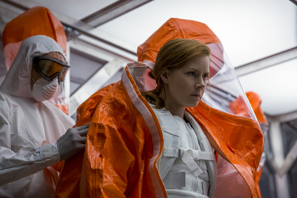 A person in a white hazmat suit helps a red-haired woman don an orange protective suit.