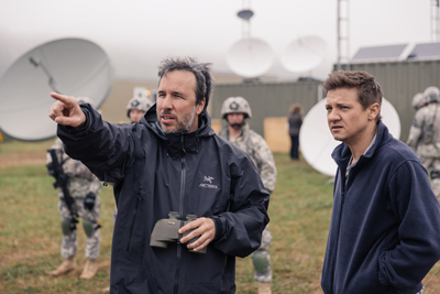 A man in a black jacket points toward the distance while another man watches him in a field.