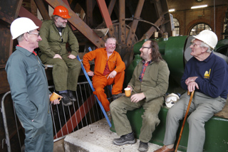 Six men in workwear and hard hats talk near large, dark metal industrial machinery and gears.