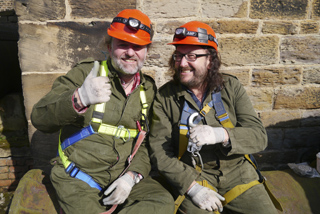 Two men in olive jumpsuits, orange hard hats, and safety harnesses sitting in front of a stone wall.