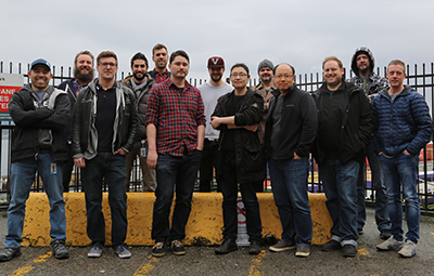A group of ten men standing in a line outdoors against a metal fence and overcast sky.