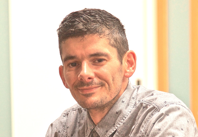 A man with short dark hair and light stubble smiling while wearing a grey patterned button-down shirt.