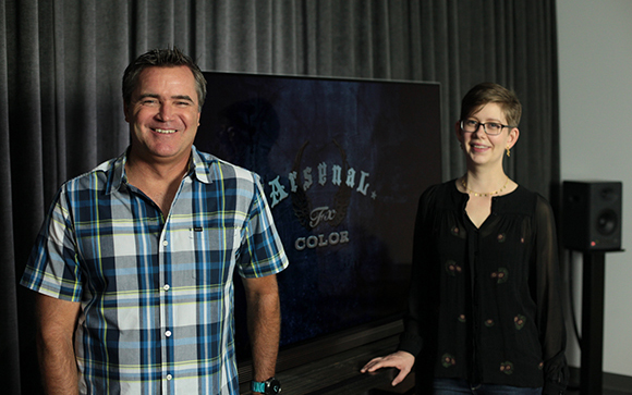 A man in a plaid shirt and a woman in a black blouse stand beside a large screen displaying 'Arsenal'.