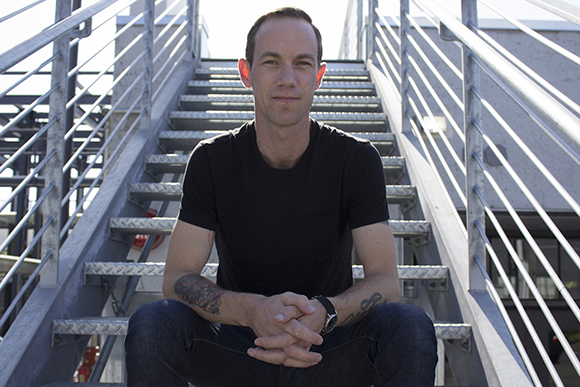A man in a black t-shirt and jeans sits on metal stairs with tattoos on his arms.