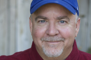 A smiling man with a grey goatee wearing a blue baseball cap and a red shirt.