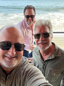 Three men wearing sunglasses smiling for a selfie outdoors near the ocean waves.