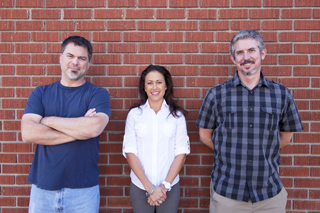 A man in a blue shirt, a woman in a white shirt, and a man in a plaid shirt stand before a red brick wall.
