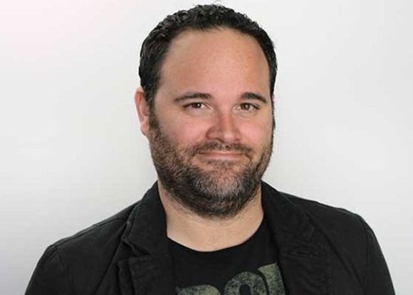 A man with dark hair and a beard wearing a black jacket and t-shirt against a white background.