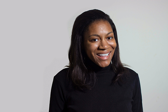 A smiling woman with dark hair wearing a black turtleneck against a light gray background.